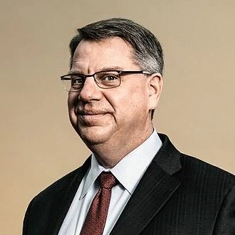 A middle-aged man with glasses, short dark hair, and a slight smile, dressed in a black suit, white shirt, and red tie, against a plain beige background.
