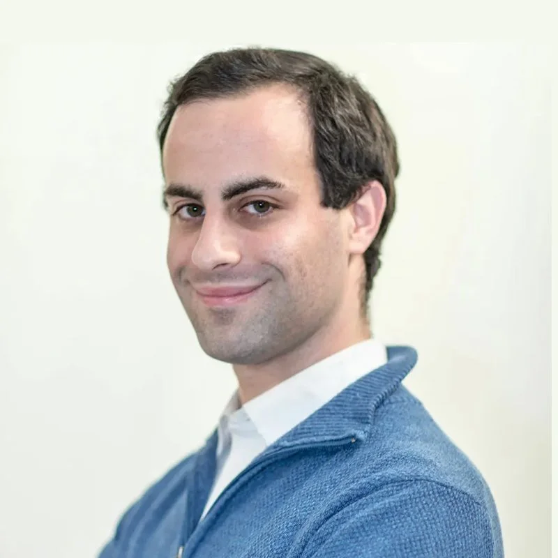 A young man smiling, wearing a blue sweater over a white collared shirt, with short dark hair and a light complexion. The background is plain and off-white.