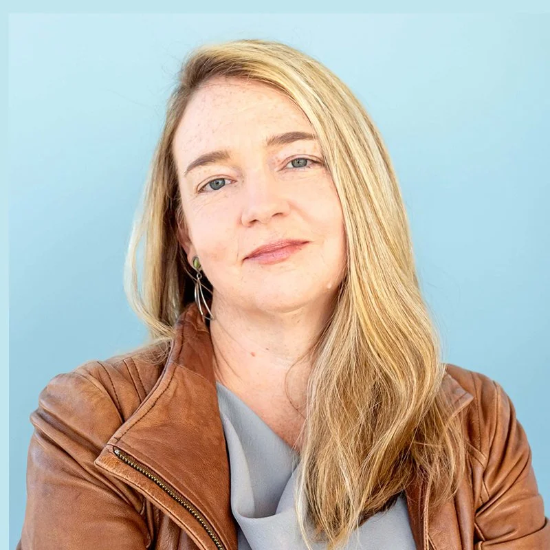 Portrait of a woman with long blonde hair, wearing a light gray top and a brown leather jacket, against a light blue background.