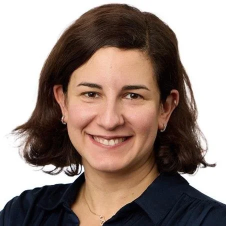 A woman with shoulder-length brown hair smiling, wearing a dark collared shirt and small earrings against a white background.