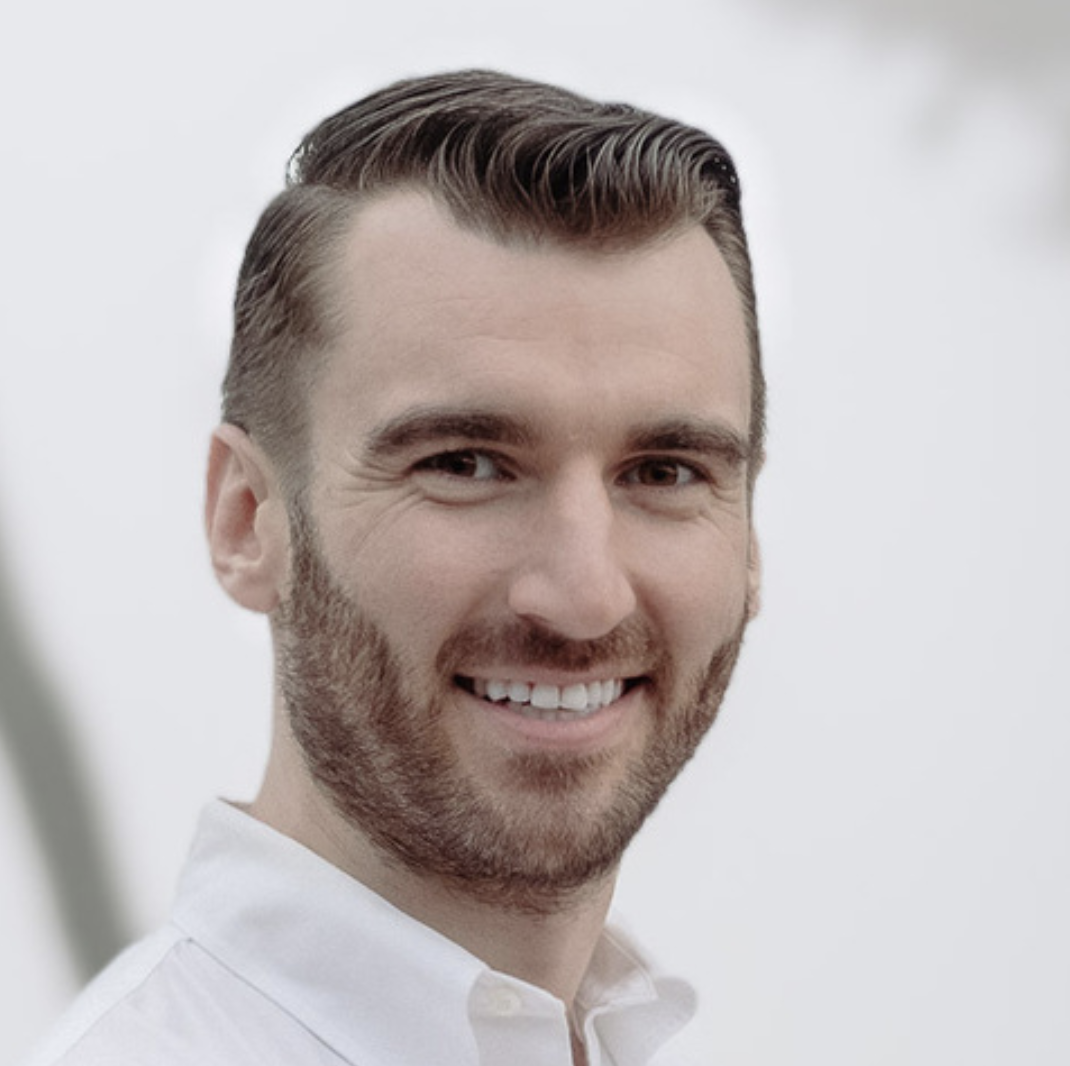 Close-up of a smiling man with short, styled hair and a beard, wearing a white shirt, against a neutral background.