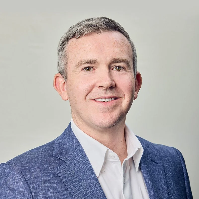 A portrait of a smiling man with short gray hair, wearing a white shirt and blue blazer, against a plain background.