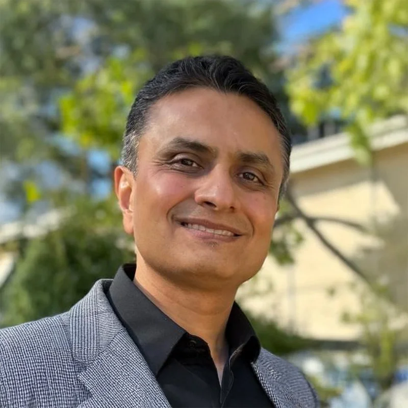 A man in a gray blazer and black shirt smiling outdoors with green trees and a building in the background.