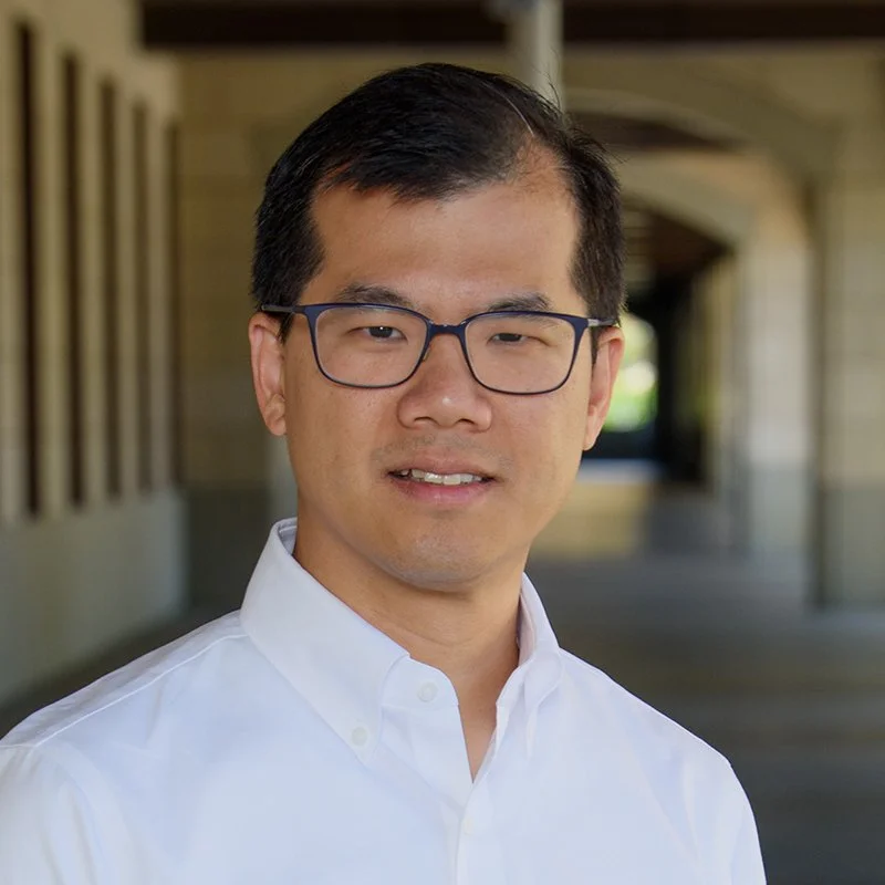 Portrait of a man with short dark hair, wearing glasses and a white collared shirt, standing outdoors in a covered walkway.