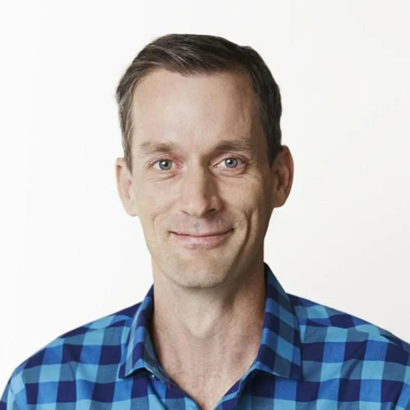 A man with short brown hair, blue eyes, wearing a blue plaid button-up shirt, smiling slightly, against a plain white background.