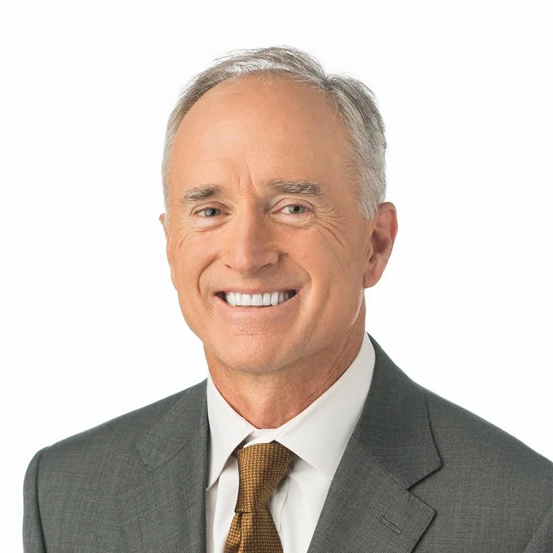 Headshot of a middle-aged man with gray hair smiling, dressed in a dark suit, white shirt, and brown tie, against a plain white background.