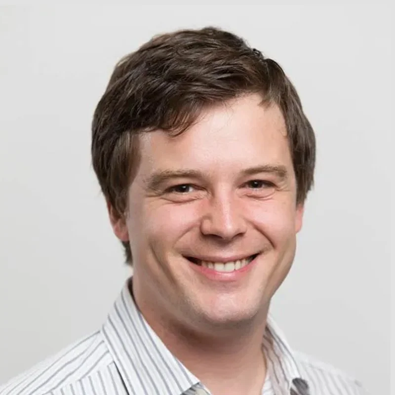 Smiling man with brown hair wearing a white and gray striped shirt.