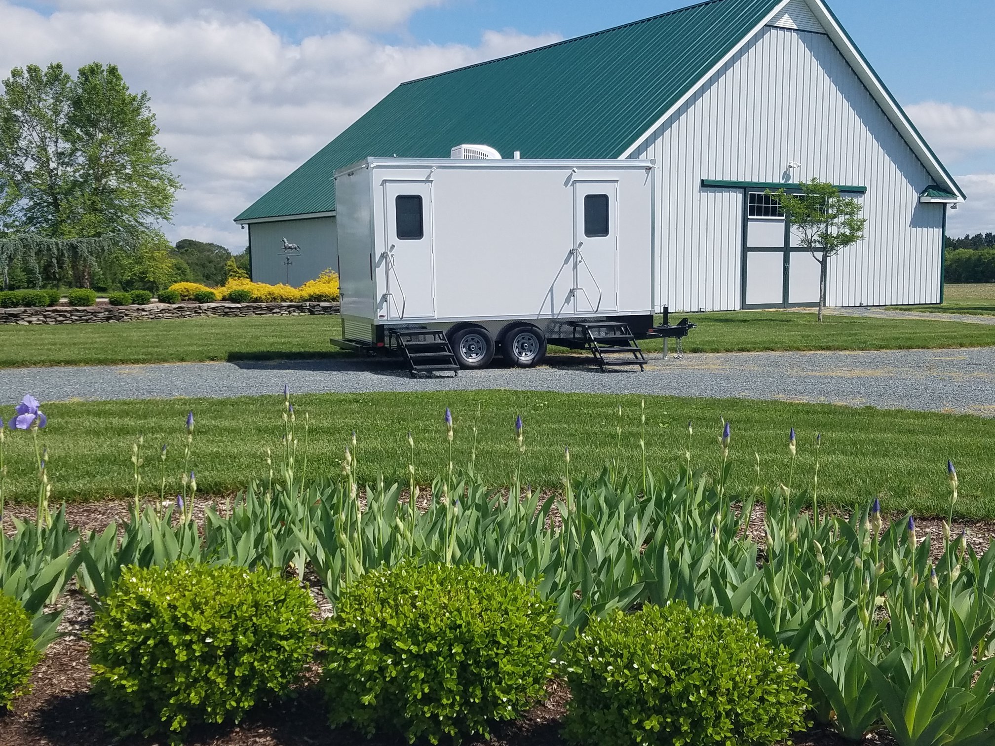 Portable restroom trailer parked on a grassy area near a large, green-roofed barn with landscaping and plants in the foreground.