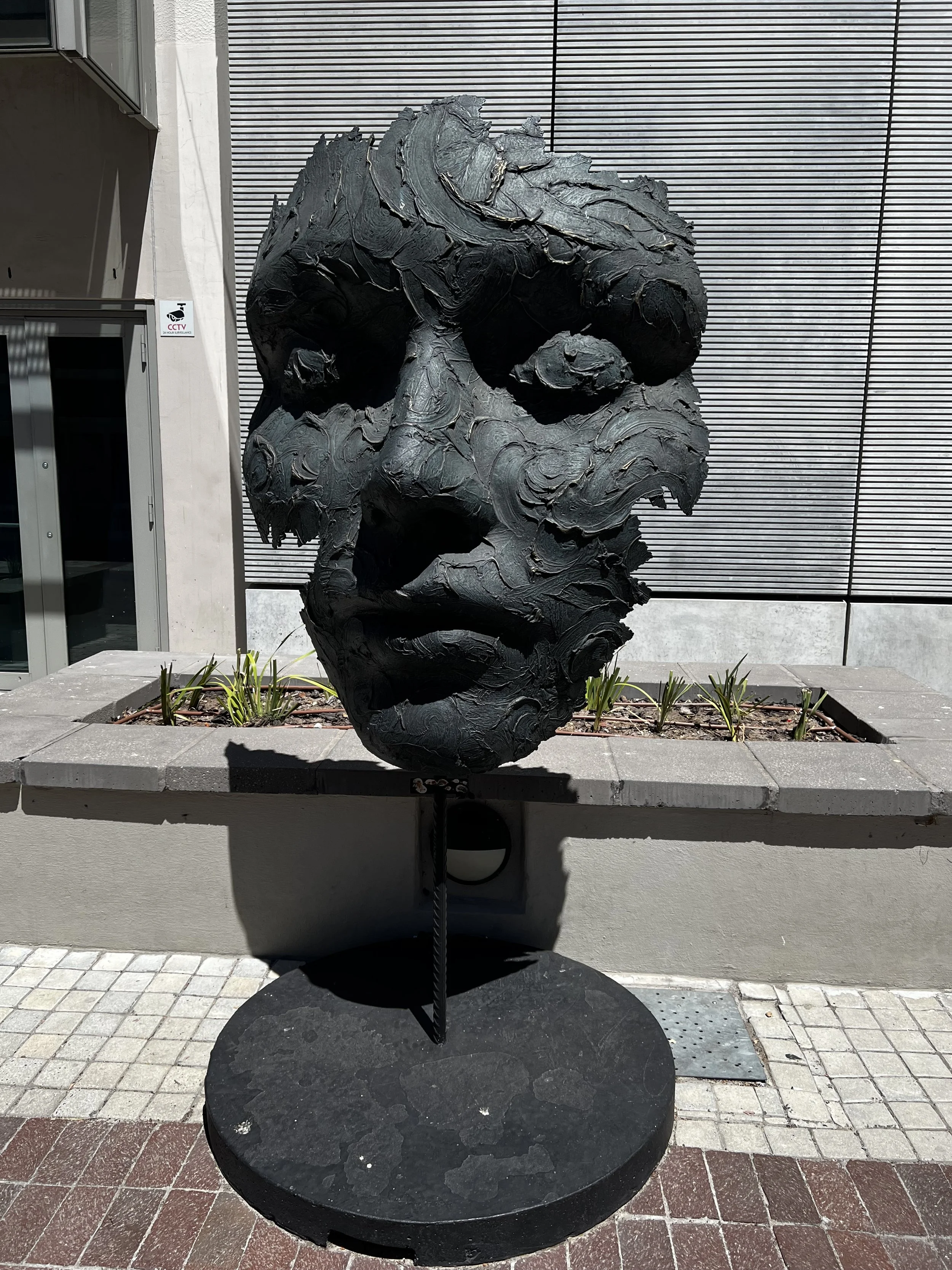 A black abstract sculpture with swirling and layered textures, positioned outdoors against a modern building with horizontal metal slats and a small garden with plants.