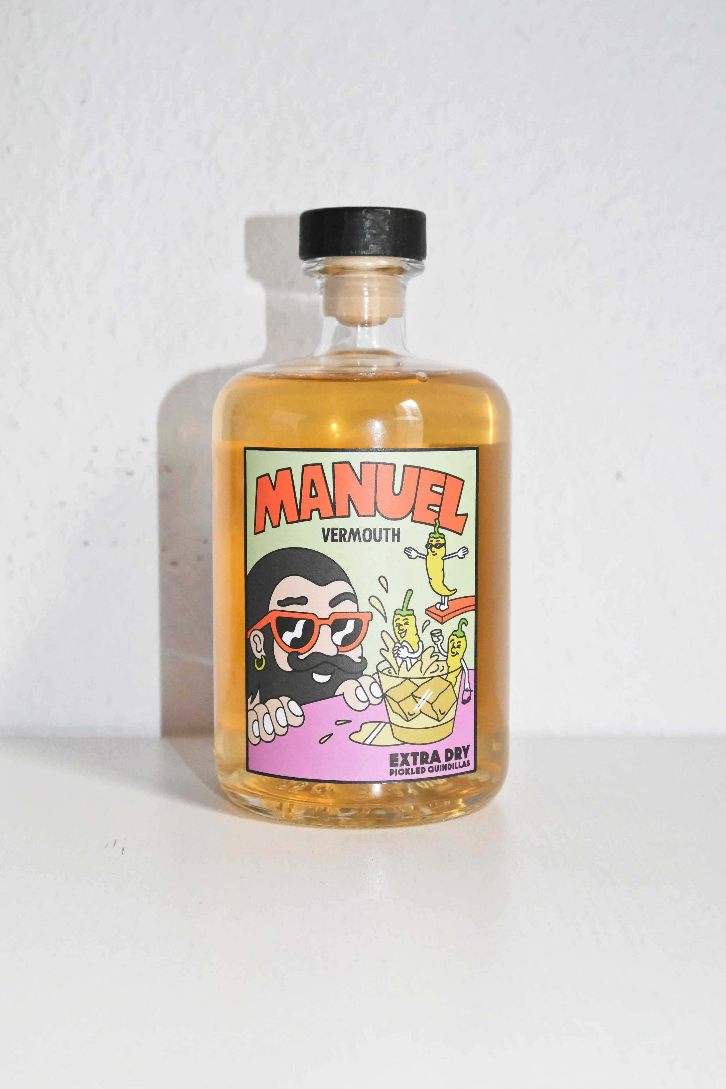 Bottle of Manuel Vermouth with cartoon label featuring a man with sunglasses and a beard, surrounded by animated pickled quindillas, with text indicating extra dry and pickled quindillas.