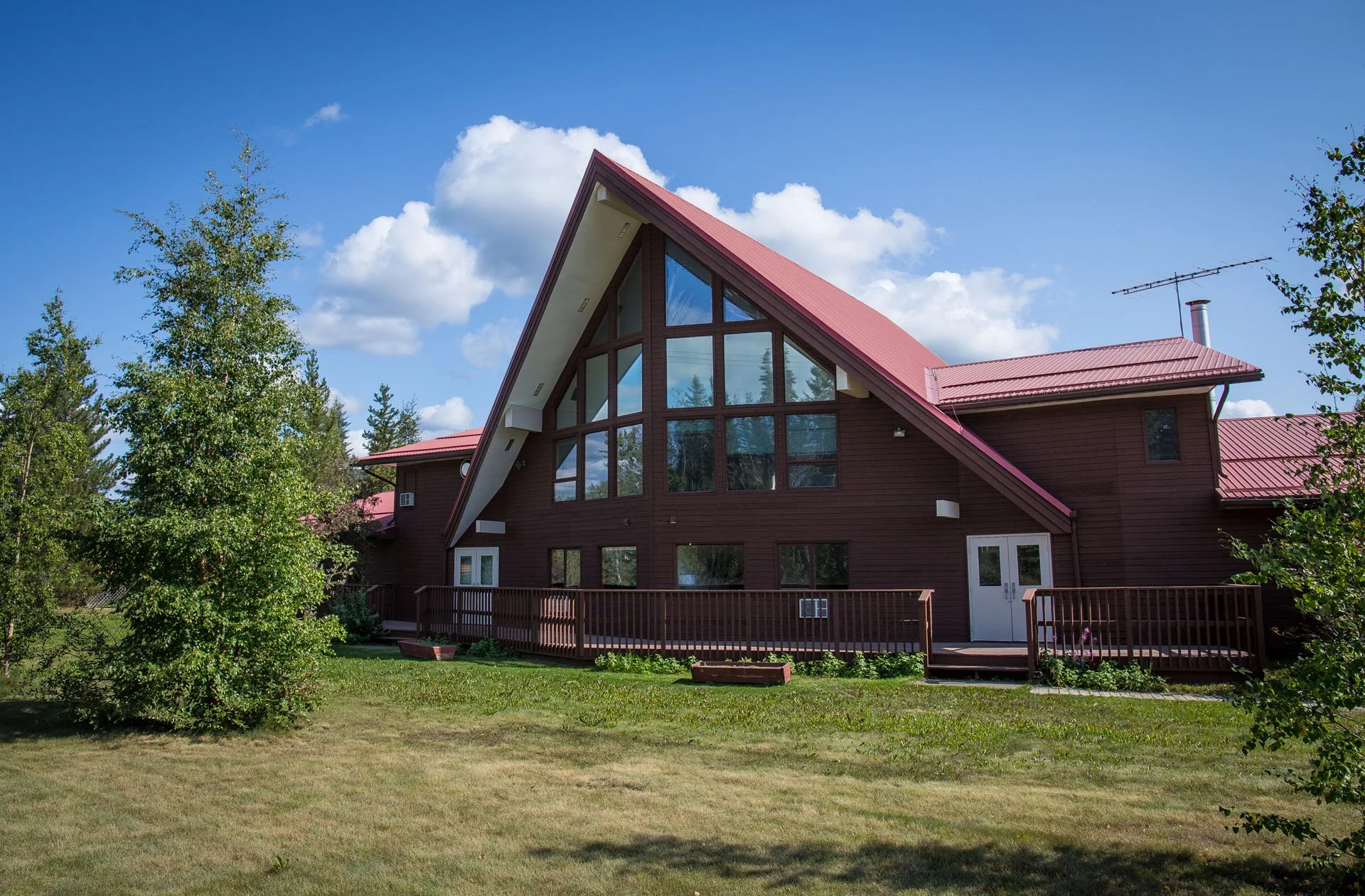 A large house with a steeply pitched roof and extensive glass windows on the front, surrounded by a grassy yard and trees under a blue sky with scattered clouds.