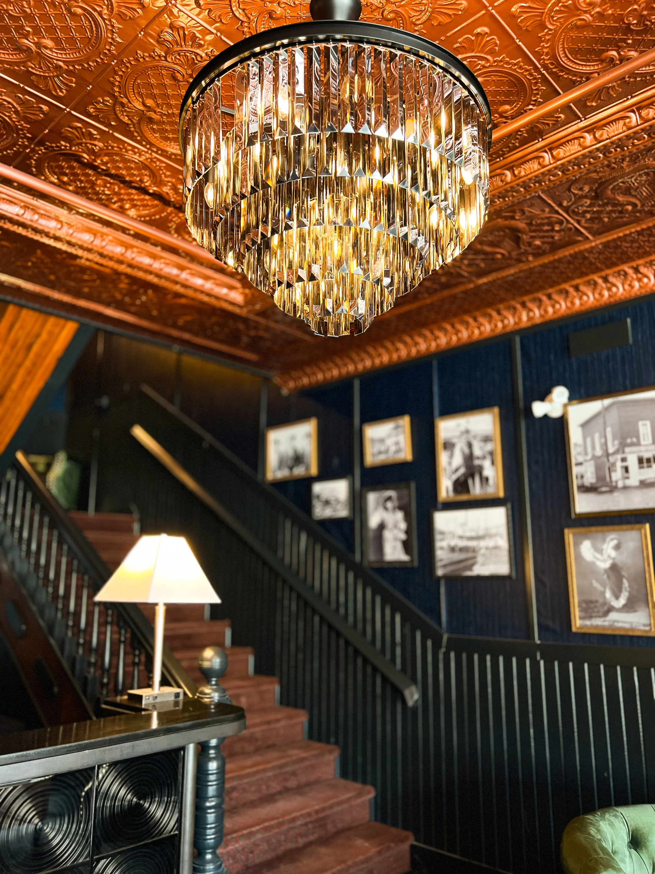 Elegant interior with a large crystal chandelier hanging from an ornate ceiling, dark walls decorated with framed black and white photographs, a staircase with wooden steps, a table with a lamp, and a green armchair in a sophisticated setting.