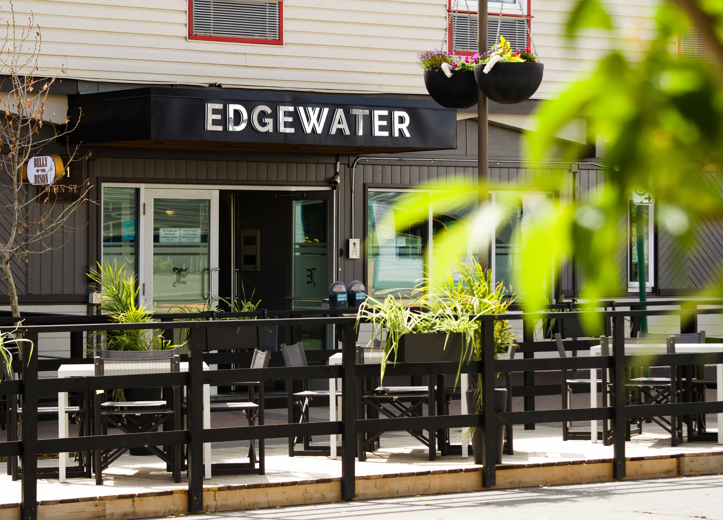 Exterior view of a restaurant named 'Edgewater' with outdoor seating, black fencing, potted plants, and a hanging flower basket outside.