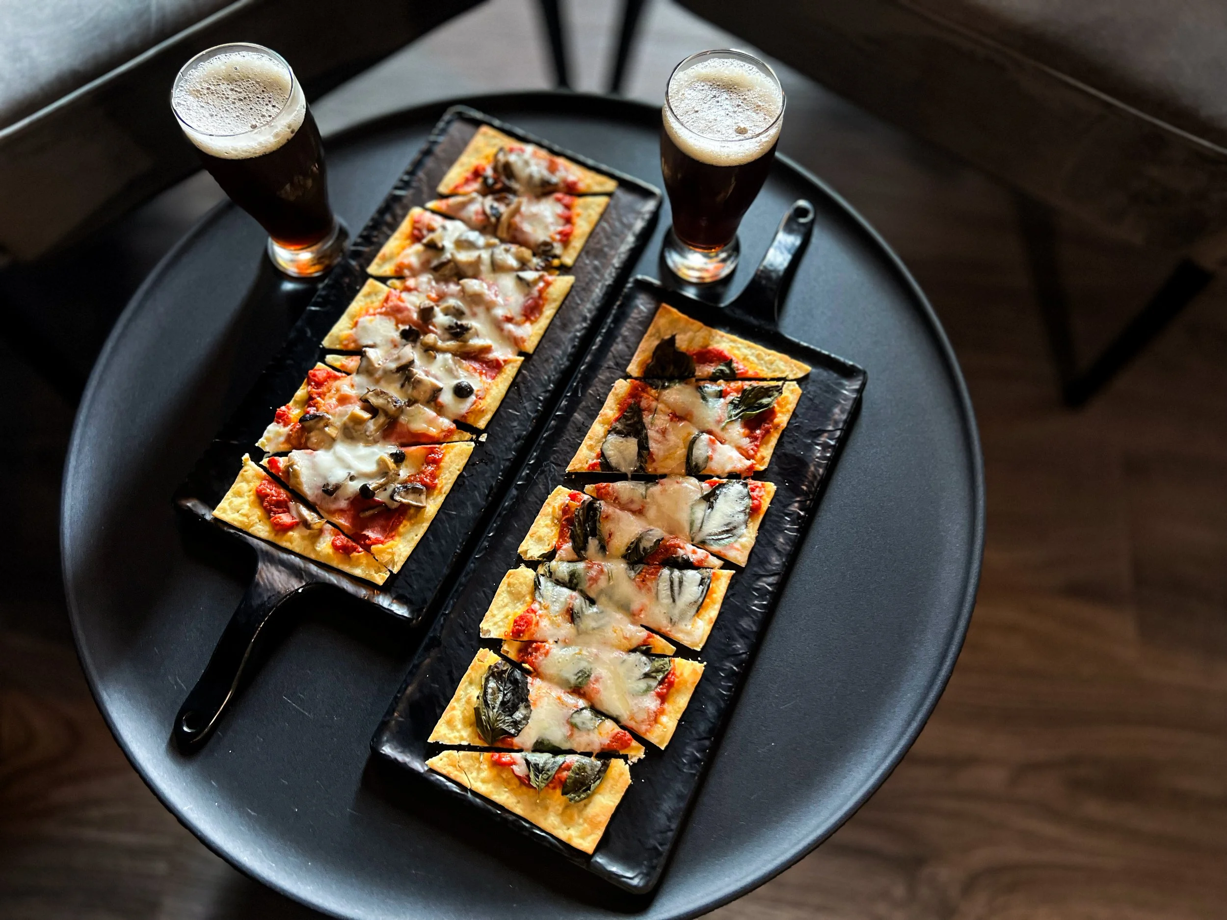 Two trays of small rectangular pizza slices with cheese, mushrooms, and basil leaves, accompanied by two glasses of dark beer on a round black table.
