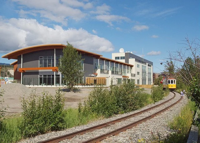 Modern building near a train track with a small yellow train passing by and trees in the foreground.