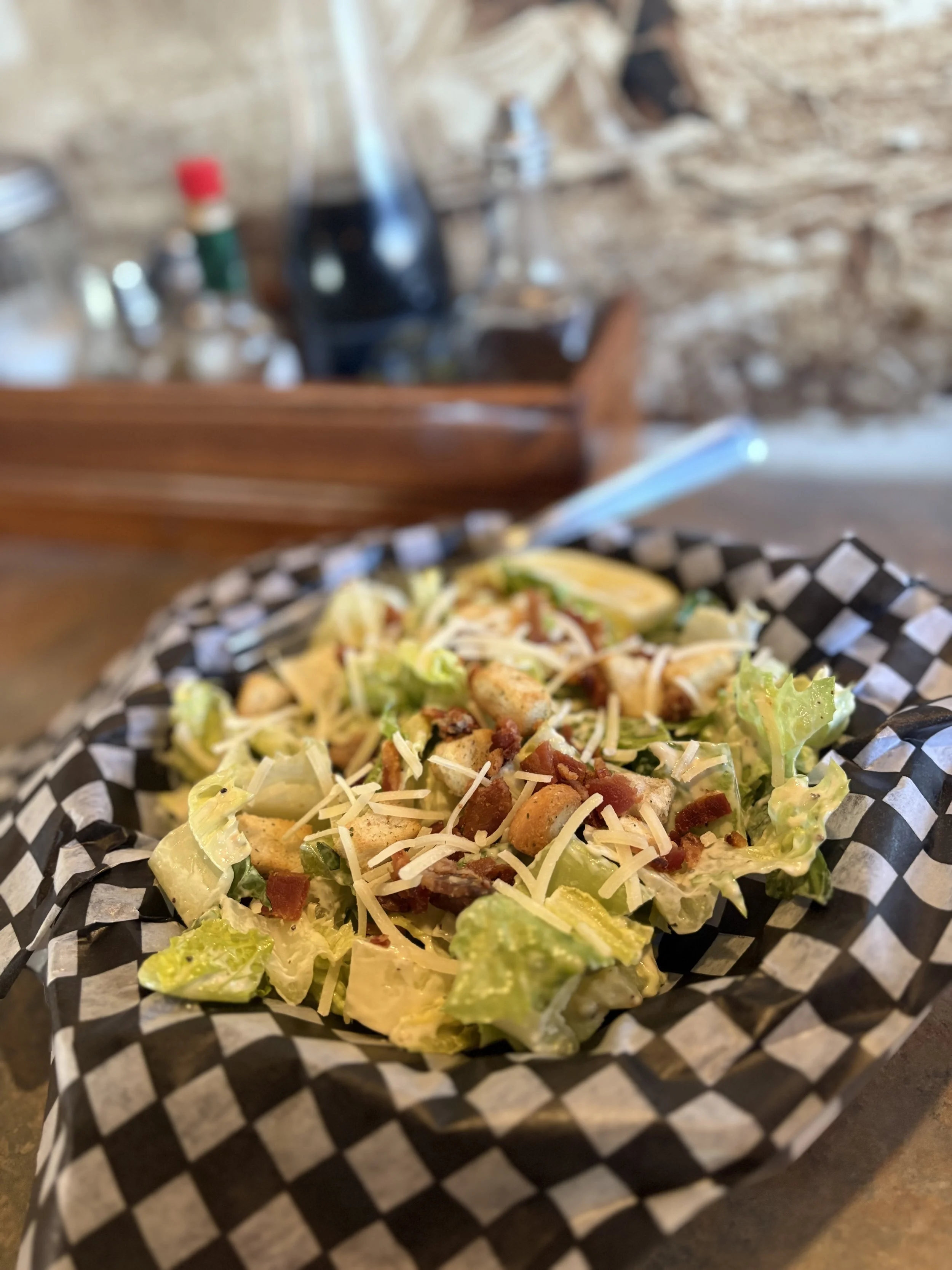 A Caesar salad with lettuce, shredded cheese, croutons, bacon bits, and lemon slices served in a checkered paper-lined basket on a wooden table.