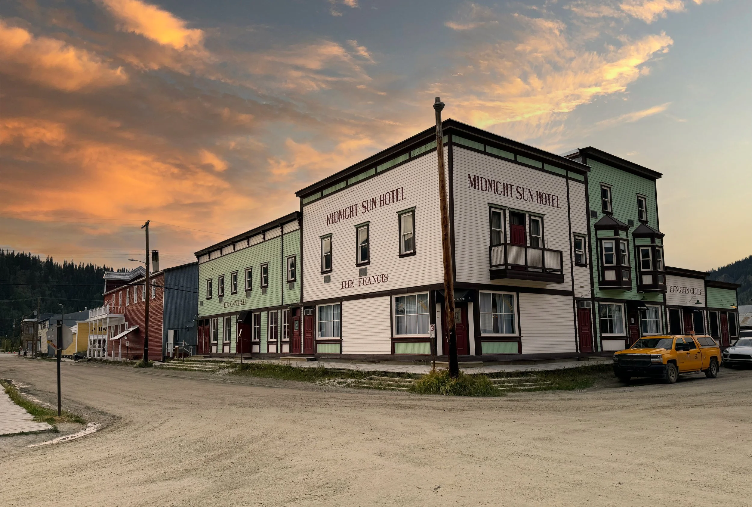 A multi-story building with signs for Midnight Sun Hotel, The Francis, and Penguin Club, under a colorful sunset sky, with parked cars and a dirt road in front.
