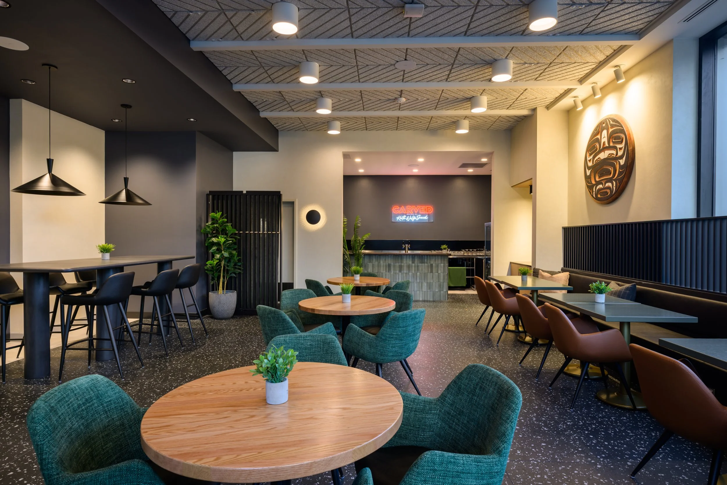 Interior of a modern restaurant or cafe with wooden tables, green and brown chairs, potted plants, and artwork on the walls. The space has a contemporary decor with black accents and a bar area in the background with a neon sign.