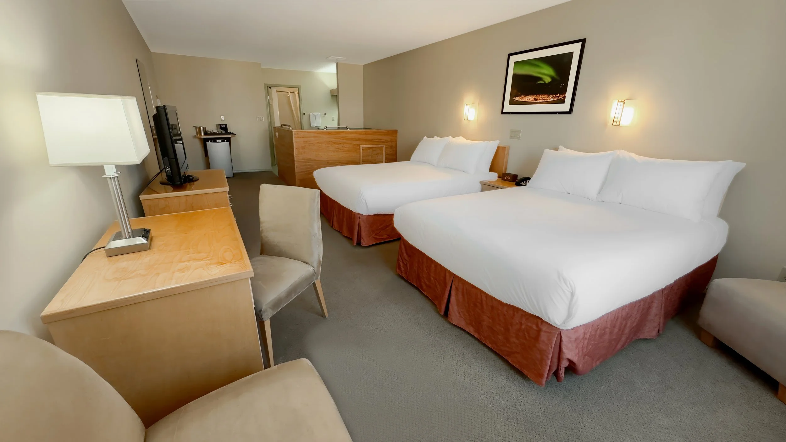 Hotel room with two queen beds, a wooden desk with lamps, television, a beige chair, and a small seating area, with artwork on the wall and a partial view of a kitchenette.