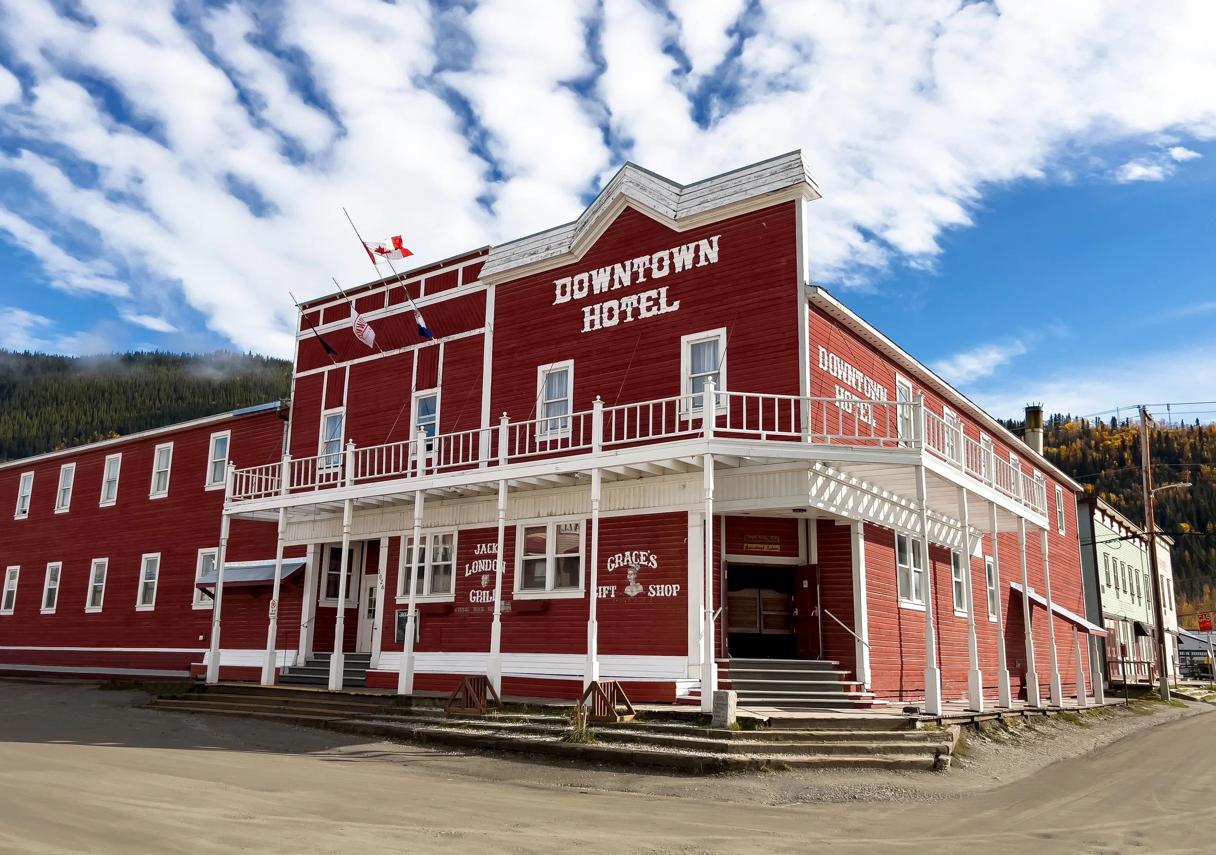 A red, two-story building labeled 'Downtown Hotel' with white trim and a balcony, set against a blue sky with clouds and a mountain with trees in the background.