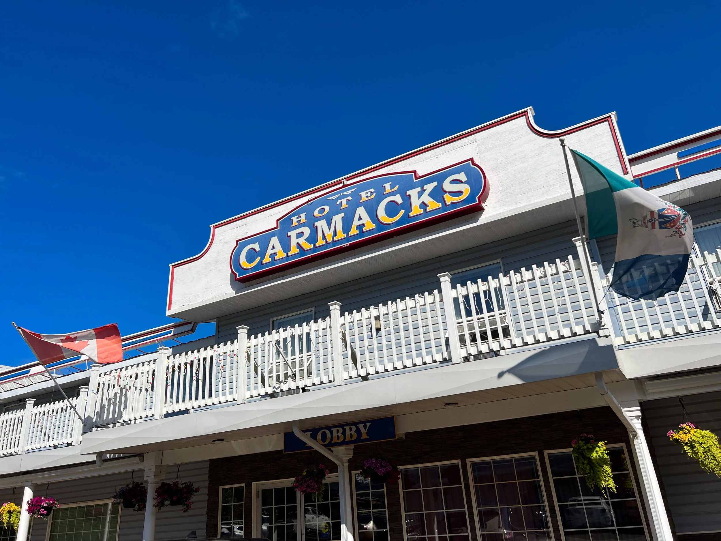 Exterior of Hotel Carmacks with a large sign, Canadian and Yukon flags, hanging flower baskets, and a sign for the lobby.