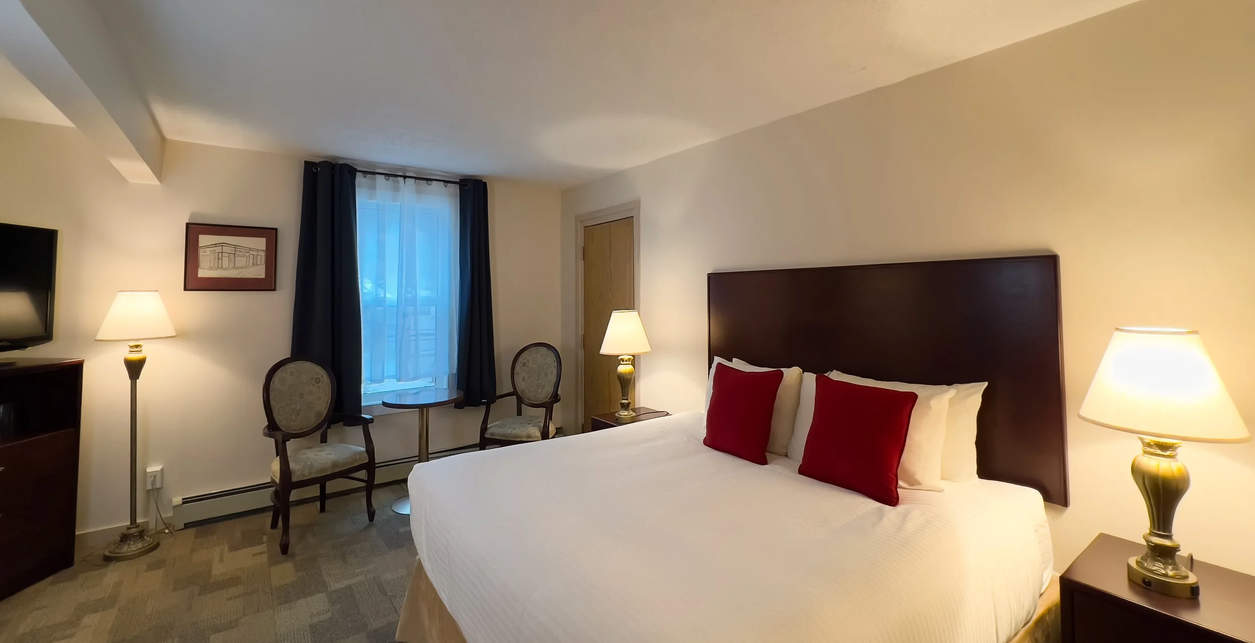 Hotel room with a large bed, dark wood headboard, beige and red pillows beside lamps, patterned carpet, window with blue curtains, two chairs with a small table, wall art, and a TV.