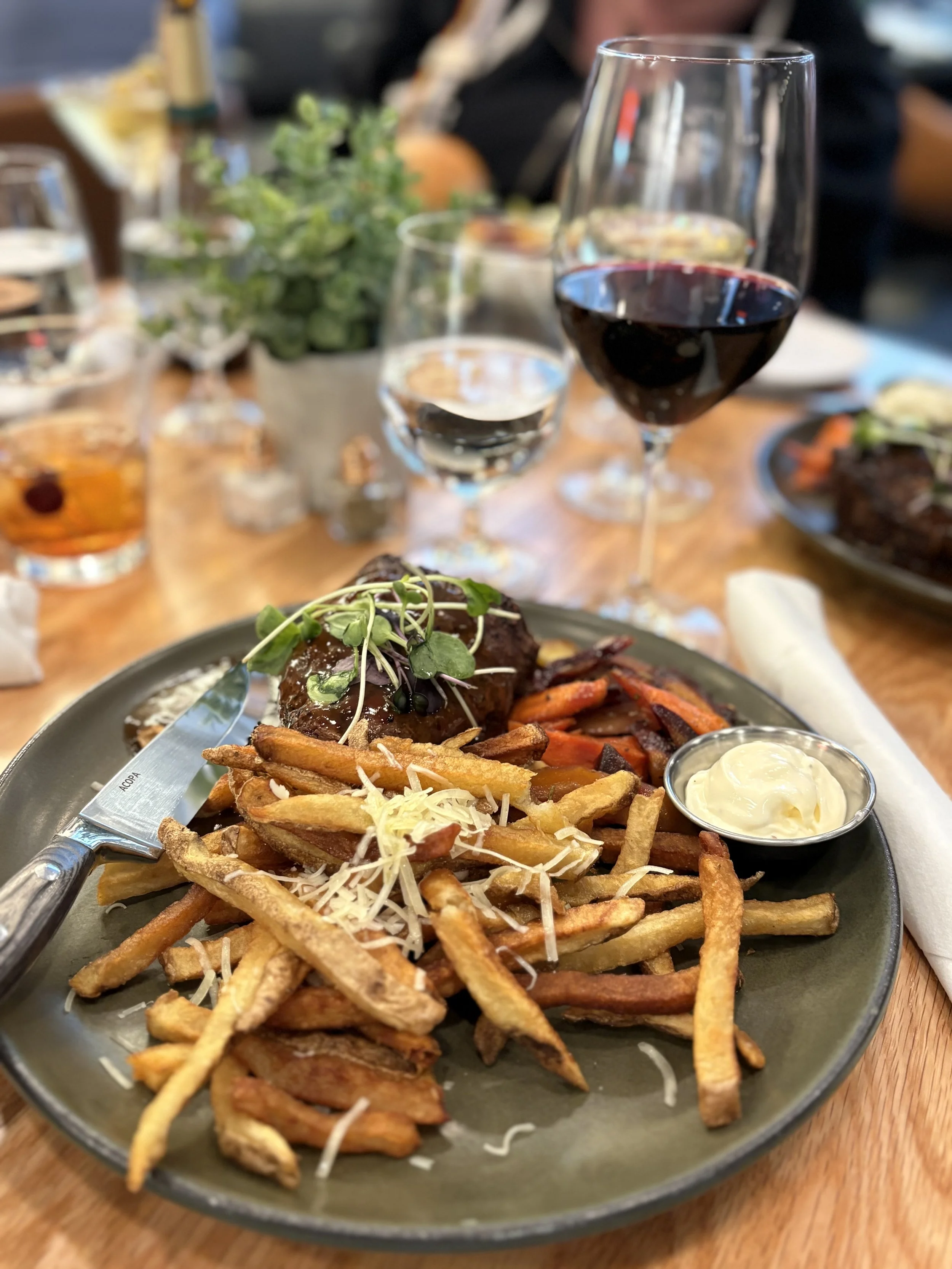 A plate of French fries with shredded cheese, a meatloaf topped with microgreens, and a side of creamy sauce, with glasses of red and white wine in the background at a restaurant.
