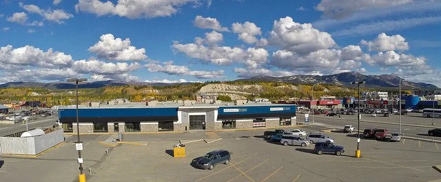 Large parking lot with several cars, a strip mall with stores, and a mountainous landscape with clouds in the background.