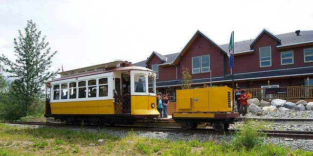 Historic narrow-gauge train with a yellow and white carriage on tracks near a red building, with people standing nearby.