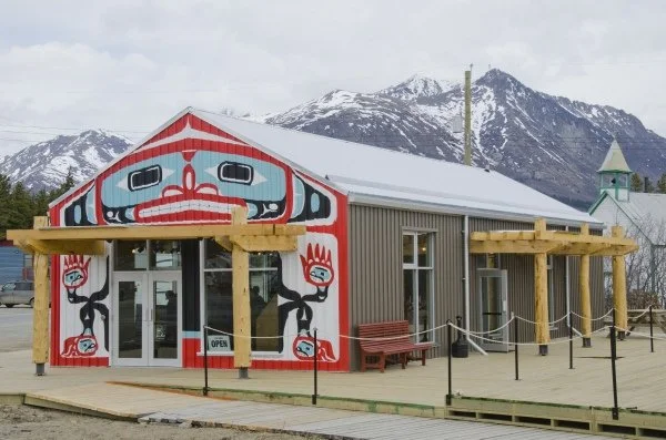 Building with a colorful totem pole design on the front, wooden railings, and a mountain range with snow in the background.