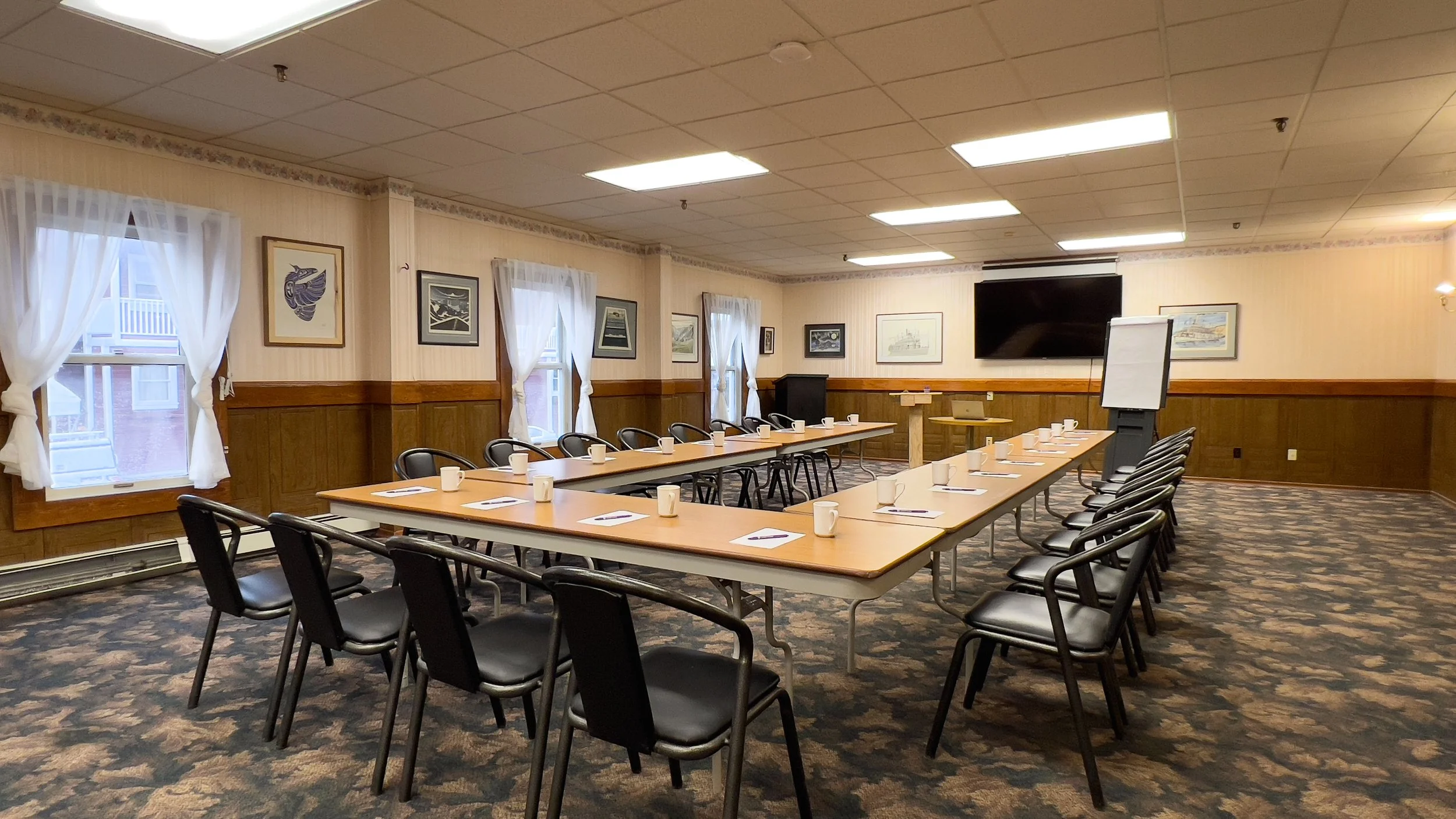 Empty conference room with U-shaped table setup, black chairs, white notepads, pens, mugs, framed artwork on walls, windows with white curtains, a large TV screen, and a whiteboard on a stand.