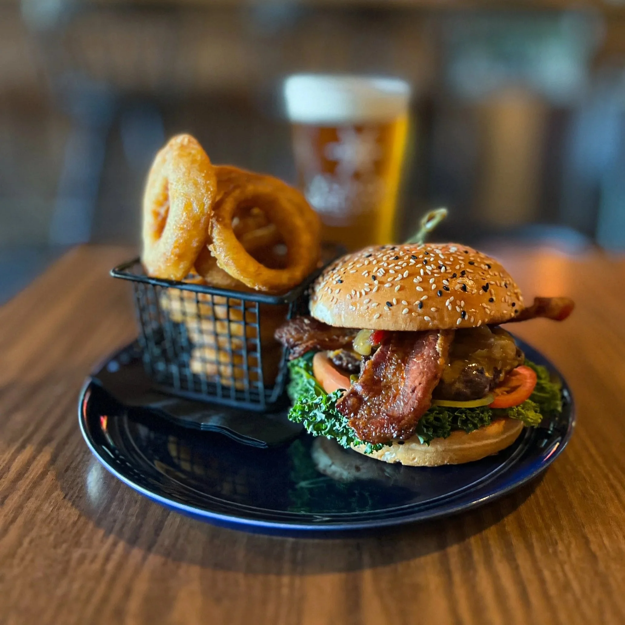 A cheeseburger with bacon, lettuce, tomato, and onion on a bun, served with onion rings and a glass of beer in the background.