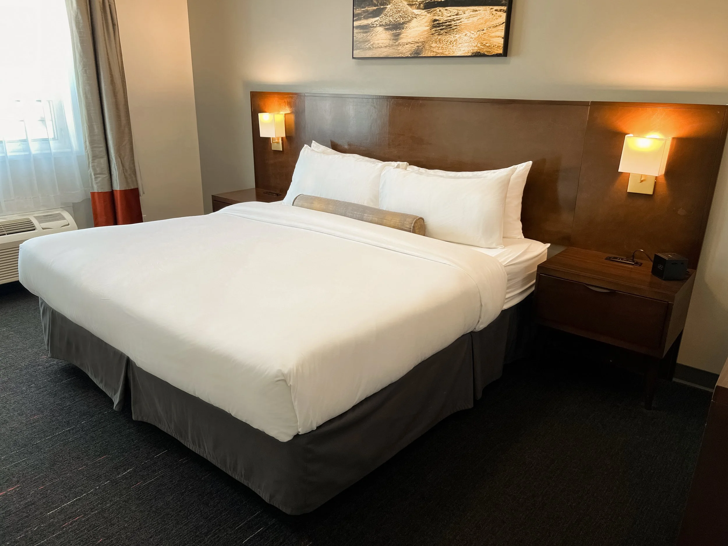 Hotel room with a large bed, white bedding, pillows, bedside tables with lamps, window with curtains, and an air conditioning unit