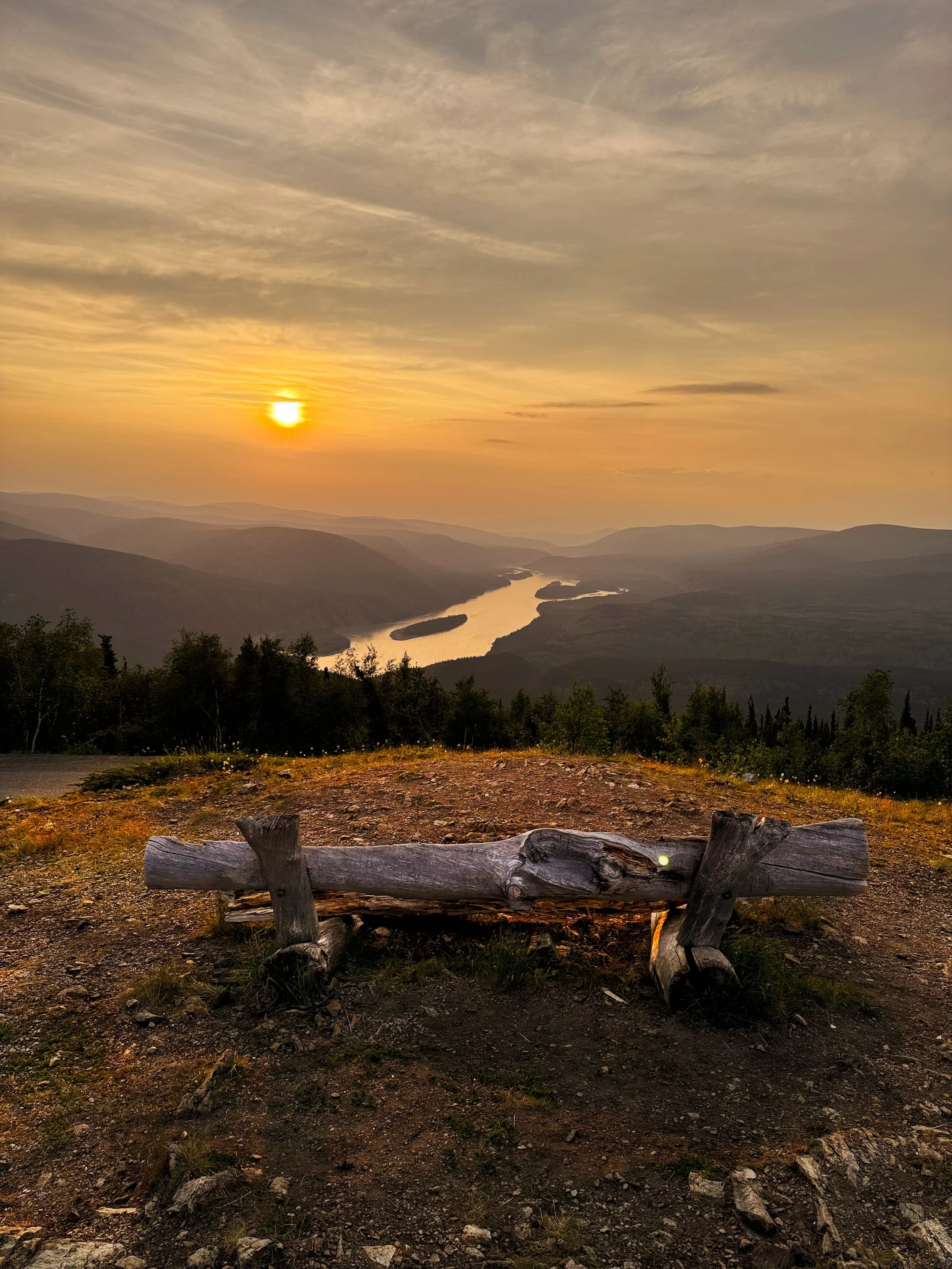 A wooden bench on a dirt ground overlooking a valley with a river, trees, and mountains during sunset with a partly cloudy sky.