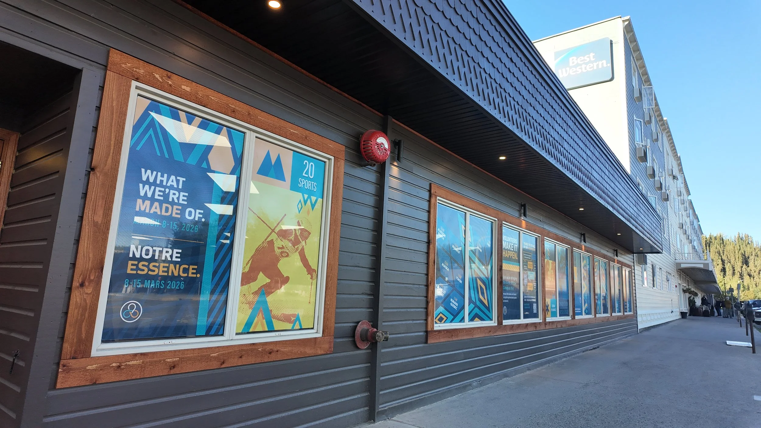 Building with blue and gray exterior, multiple posters in windows promoting sporting events and volunteer activities, Best Western hotel sign visible on taller building in the background.
