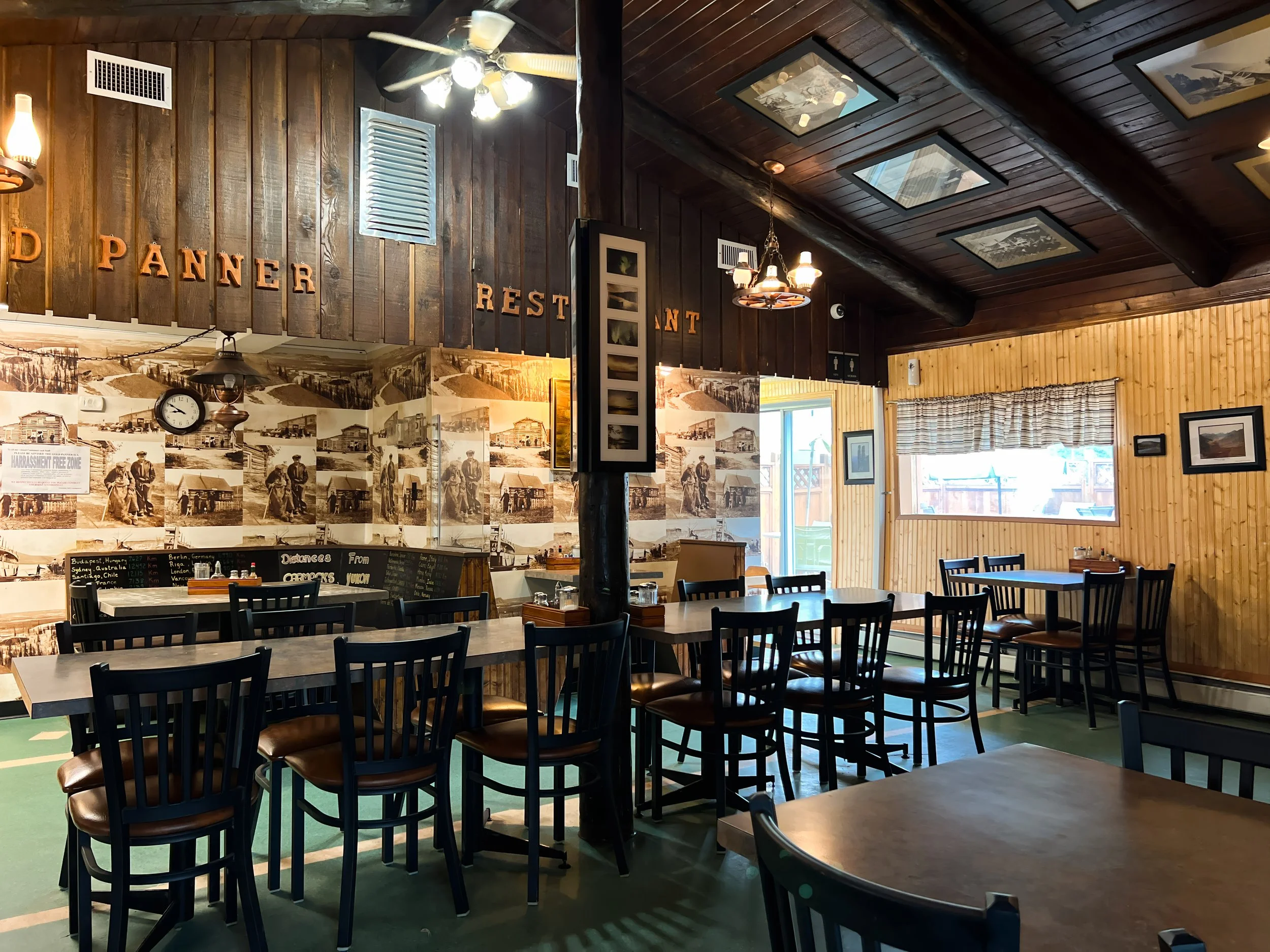 Interior of a rustic restaurant with wooden walls and ceiling, tables and chairs, vintage photos on the wall, and warm lighting.