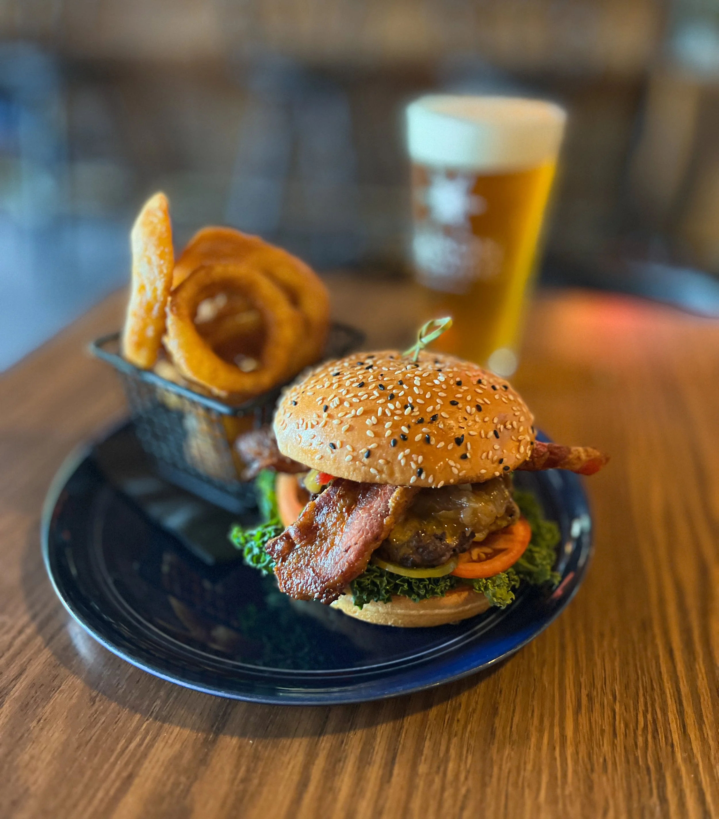 A gourmet burger with bacon, lettuce, tomato, and cheese on a sesame seed bun, served with onion rings and a glass of beer in the background.