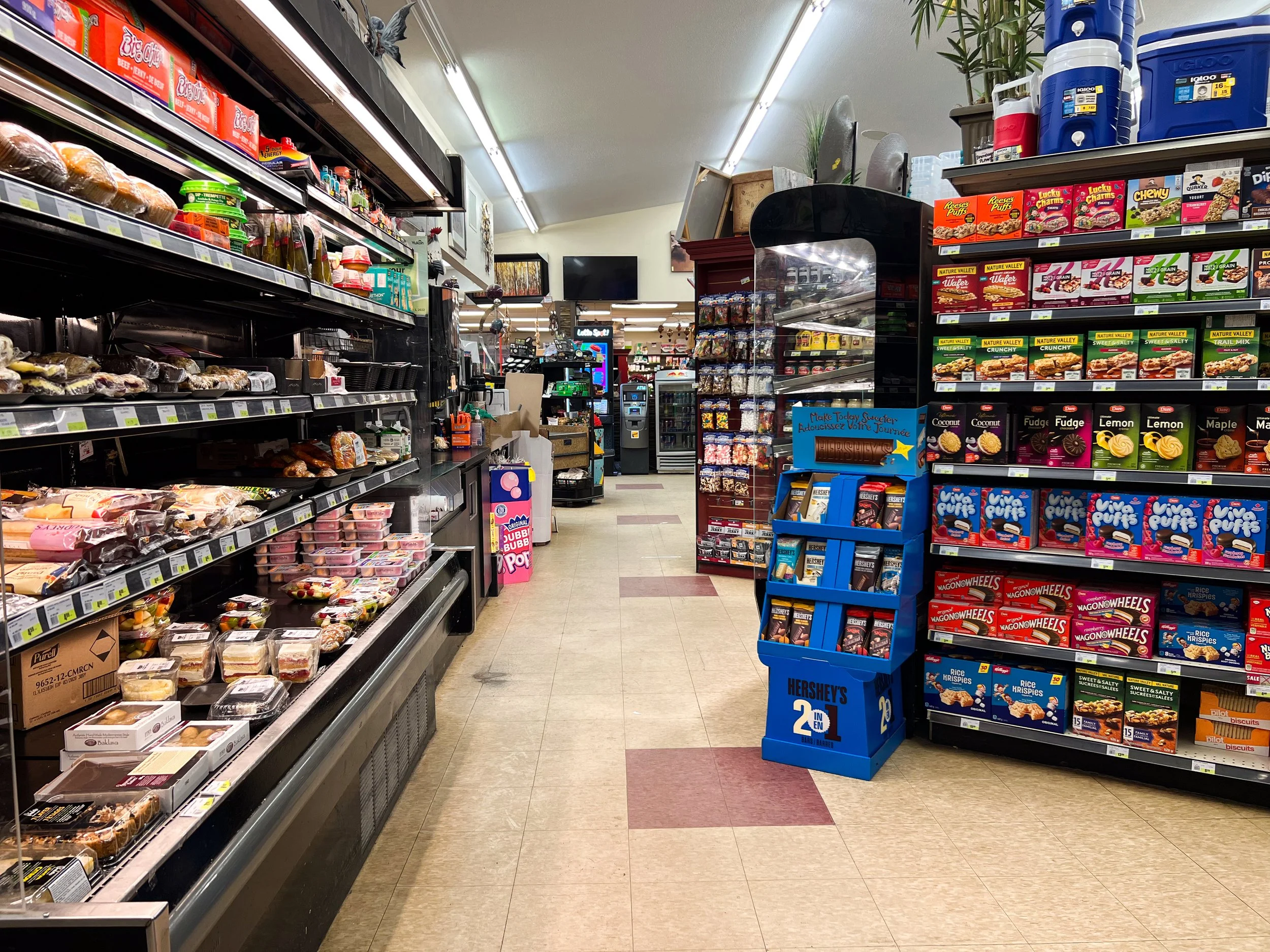 A grocery store aisle with shelves stocked with snacks, candy, and baked goods on the right side, and refrigerated items on the left. The aisle is well-lit and tidy, with a checkout area in the background.