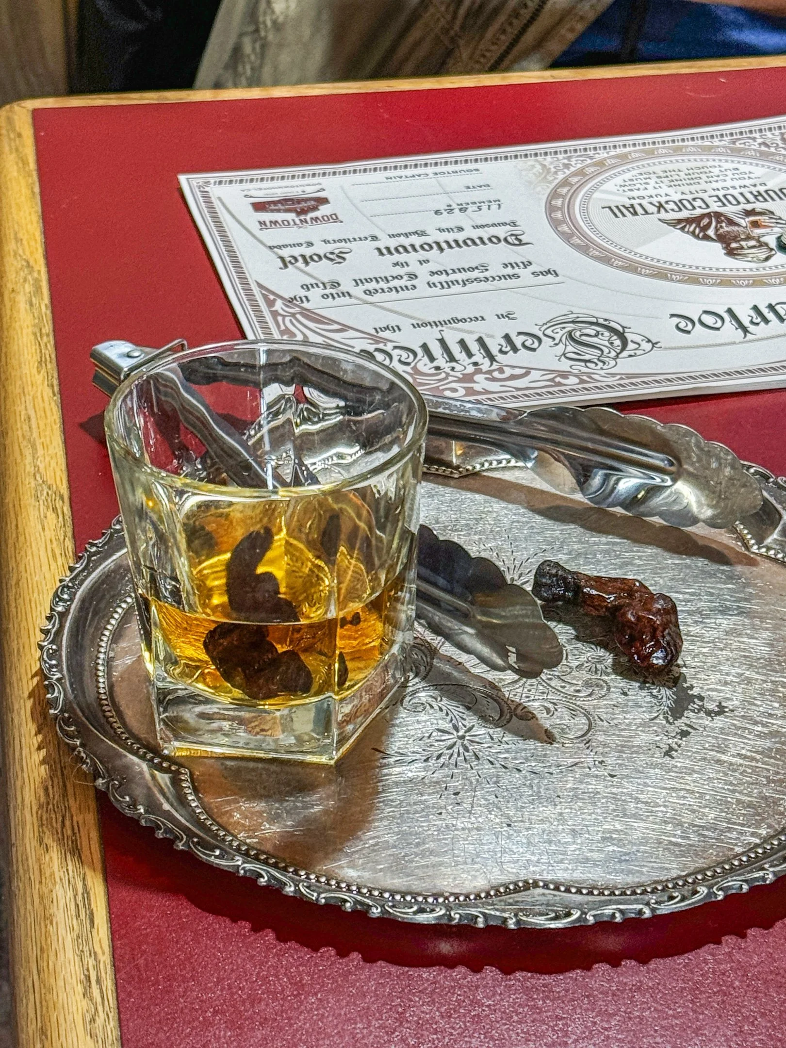 A glass of whiskey with a few dark candies inside is placed on a decorative silver tray. A pair of tongs rests on the tray. There is a red menu or placemat and a certificate or menu on top, all on a wooden table.