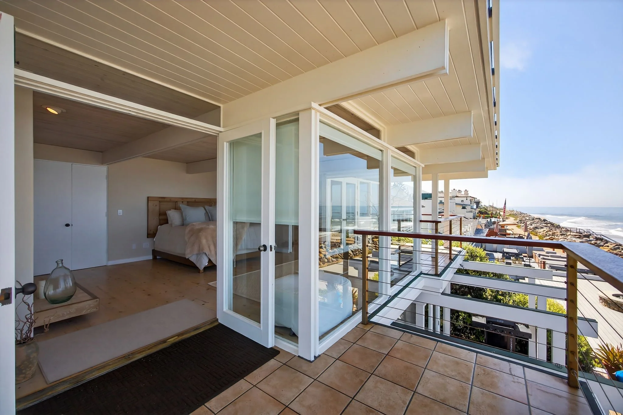 Balcony overlooking a beach with horizon and ocean waves, adjacent to a bedroom with a bed and sliding glass doors.