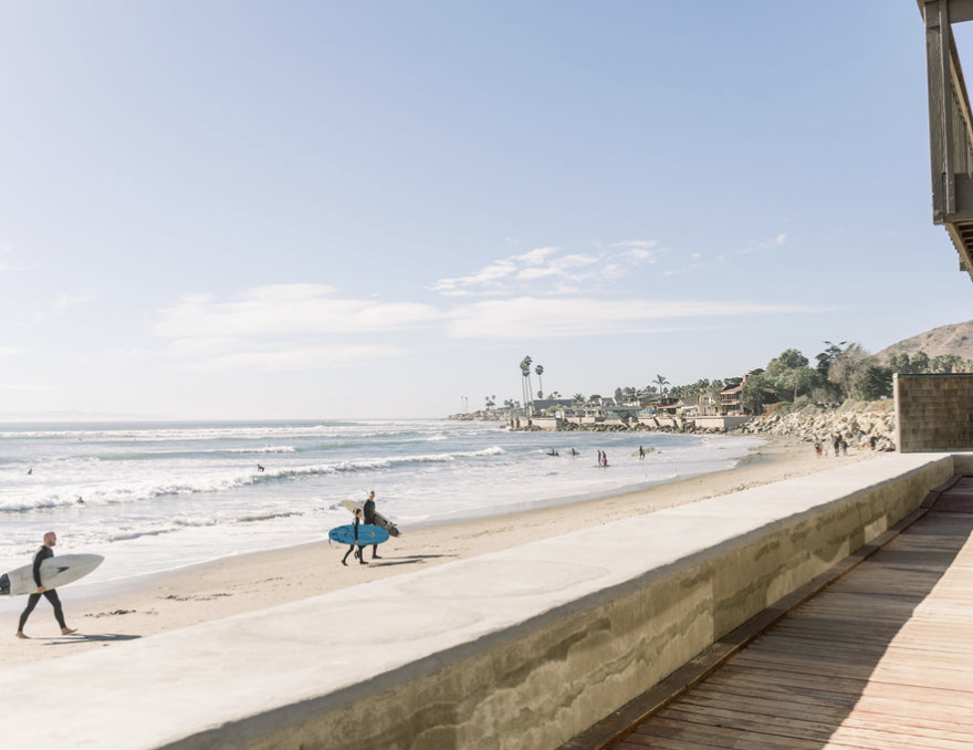 Beach scene with surfers walking along the shoreline, some carrying surfboards. The ocean has gentle waves, and a few surfers are in the water. Palm trees and residential buildings are visible in the distance under a clear sky.