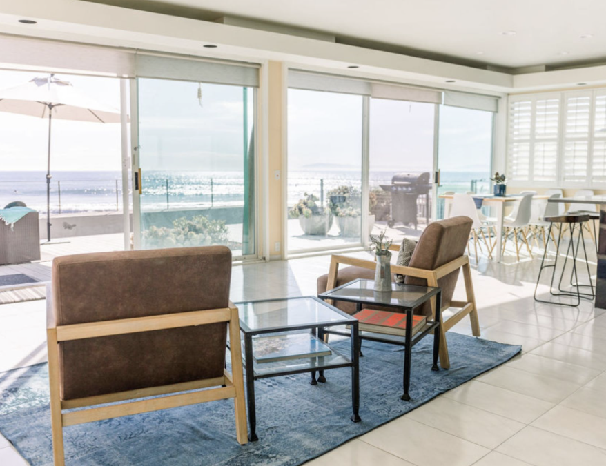 Living room with large sliding glass doors overlooking the beach, featuring two armchairs, a small glass coffee table, and a blue area rug, with a dining area and balcony outside.