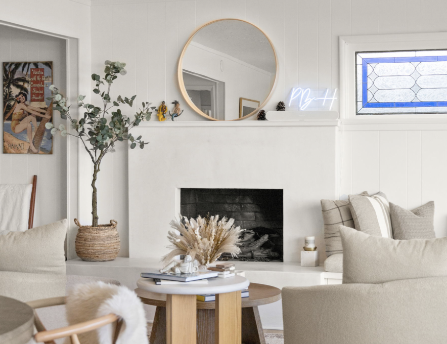 Living room with a white fireplace, round mirror, patterned plaid pillows, beige sofa, a small side table with books and dried flowers, a large potted eucalyptus plant, a Nevada vintage poster, and a stained glass window.