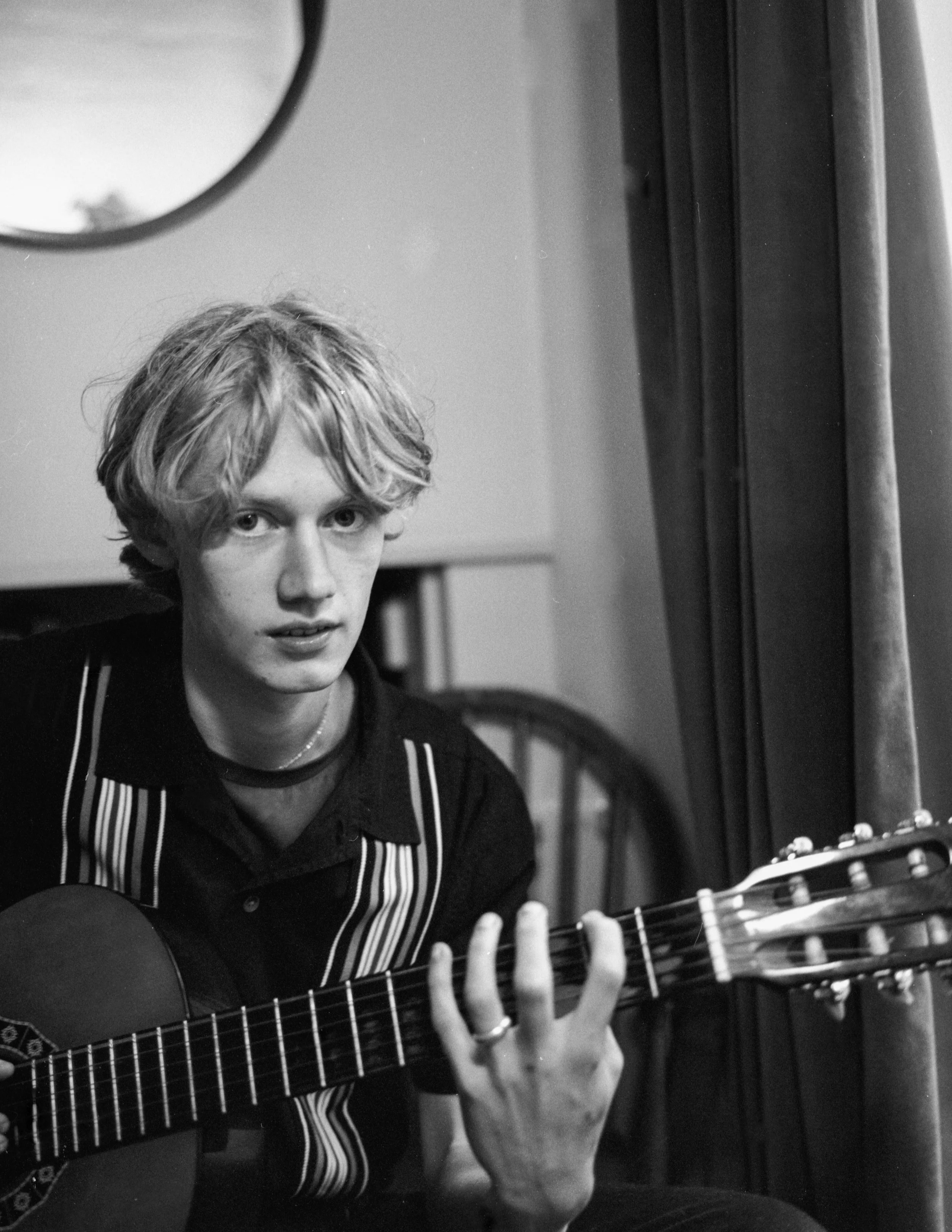 A person with wavy hair playing an acoustic guitar, wearing a striped apron, and sitting near a window with curtains, with part of their face visible in a black and white photo.