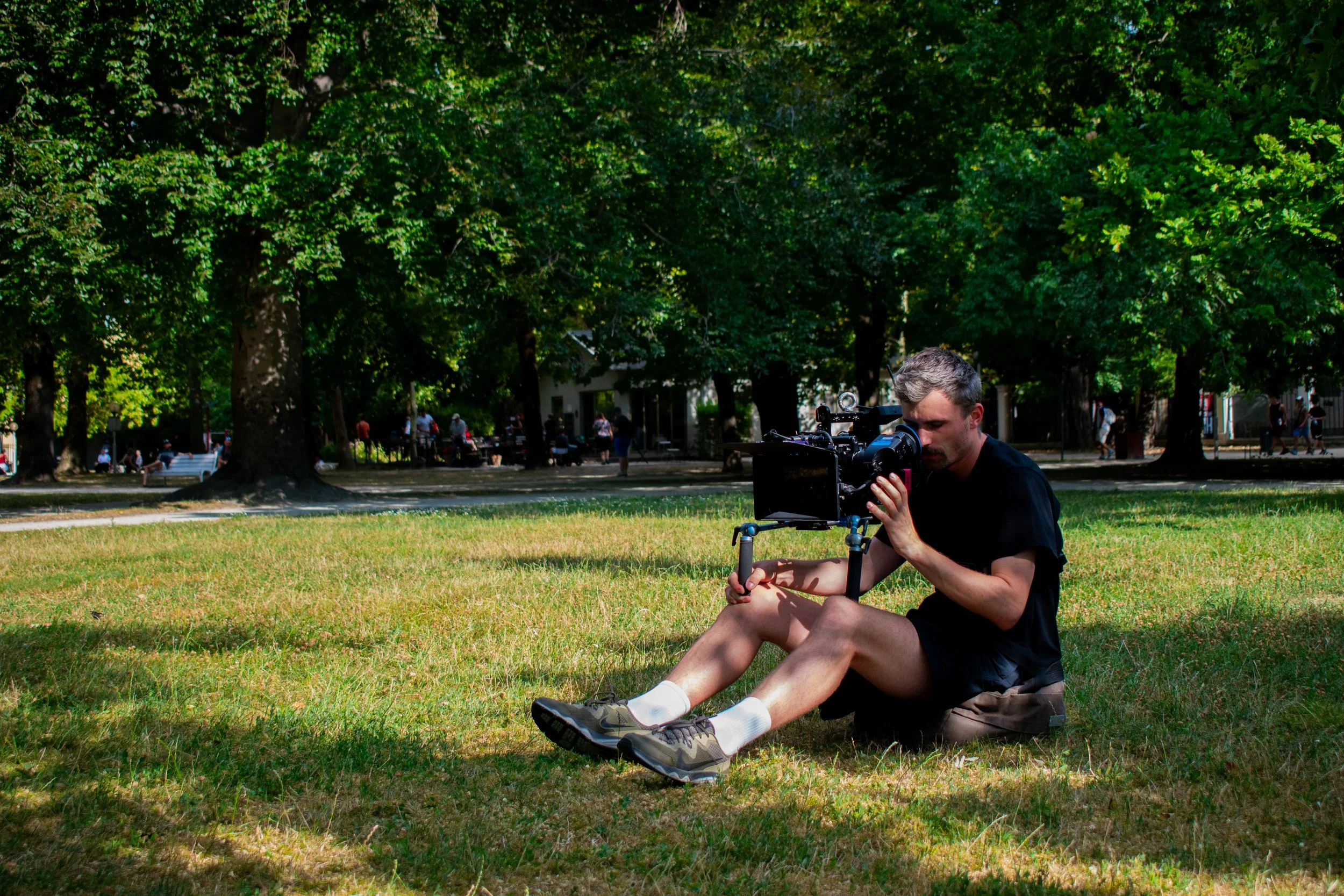 A man sitting on the grass in a park while adjusting a professional video camera on a tripod, with trees and people walking in the background on a sunny day.