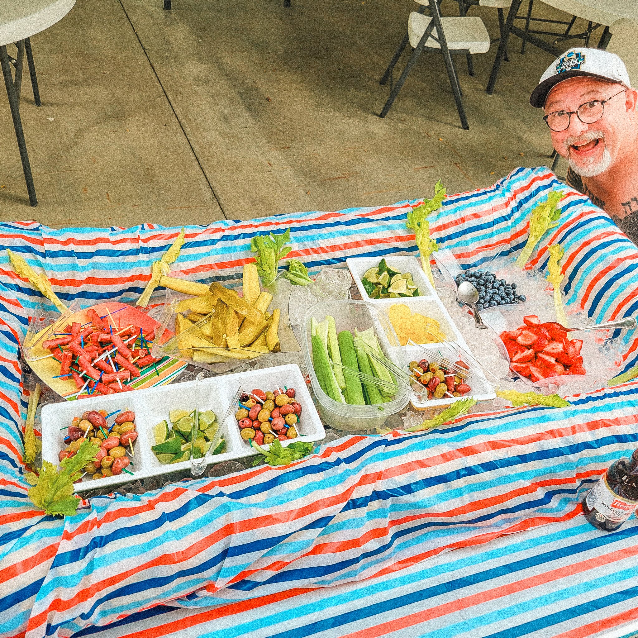 A man smiling at a festive table with various fruits, vegetables, and snacks arranged on a striped tablecloth.