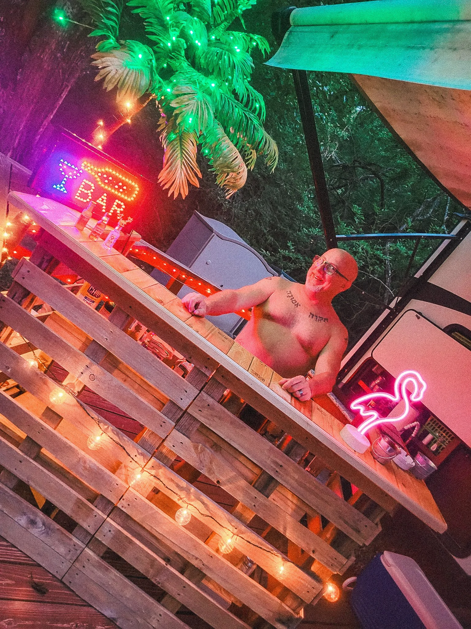 A shirtless man with glasses and tattoos standing behind a wooden bar. The scene is illuminated with colorful neon lights, including a flamingo sign and a 'Beer' sign. There are tropical plants and string lights in the background, suggesting a festiv