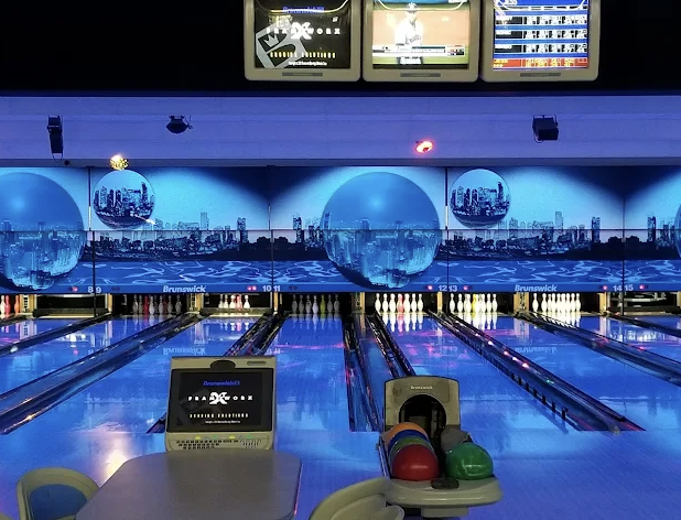 Bowling alley with multiple lanes, digital score screens, colorful bowling balls on a rack, and a cityscape aquarium mural in the background.
