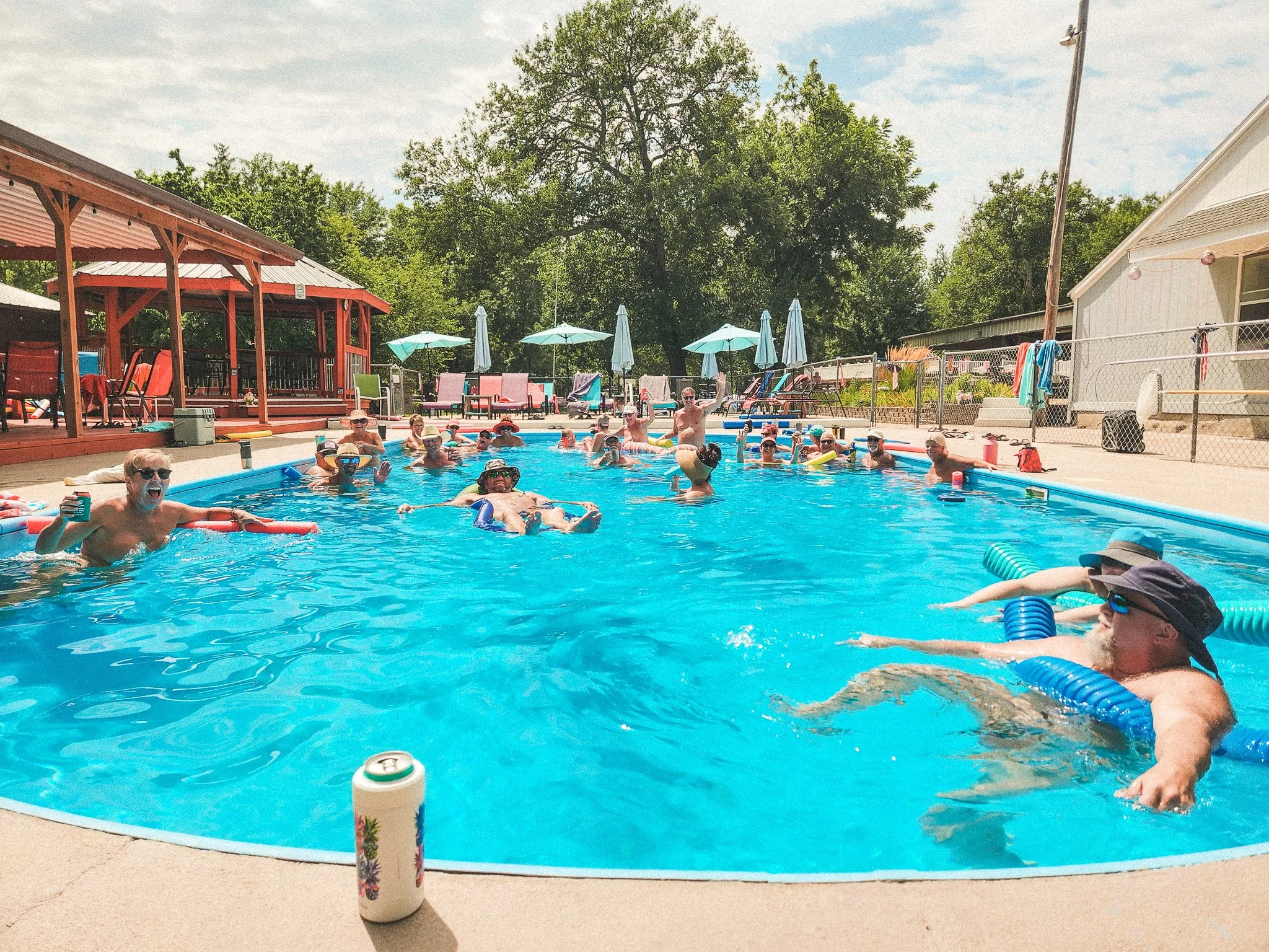 A group of people enjoying a swim and relaxing in a backyard swimming pool on a sunny day, with poolside lounge chairs, umbrellas, and a poolside pavilion in the background.
