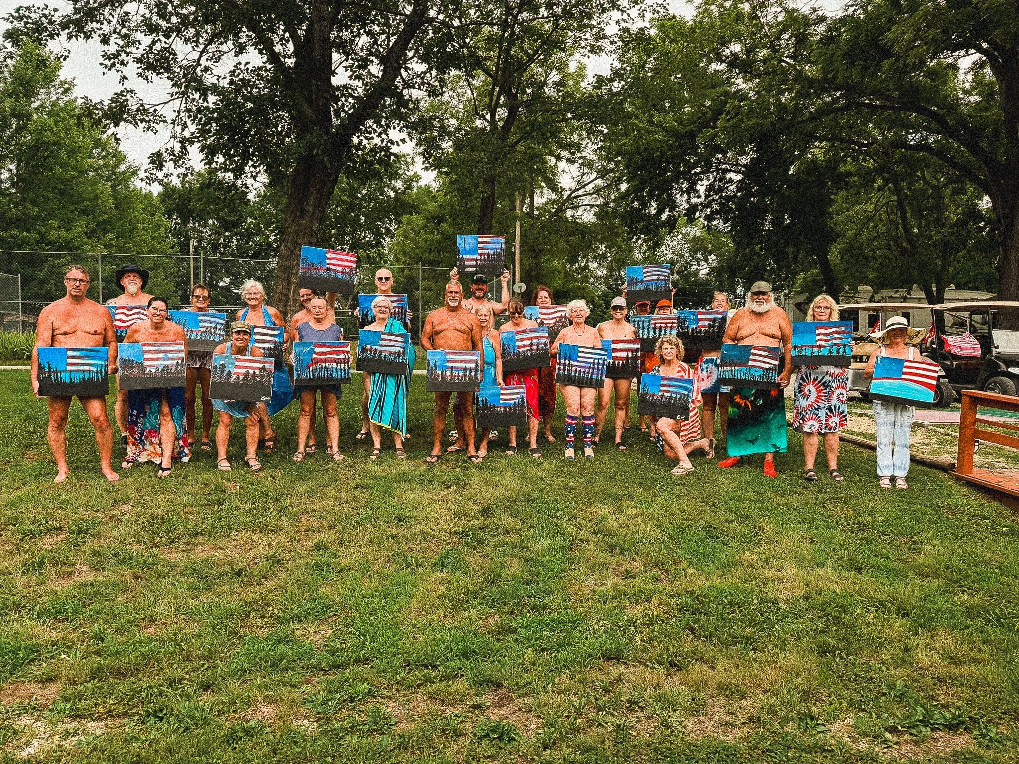 Group of people outdoors holding paintings of American flags, wearing casual summer clothing and swimsuits, under large trees on a grassy area.